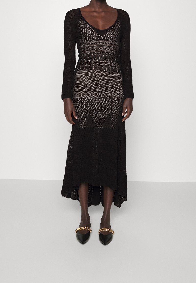 Femme portant une robe en maille noire à manches longues avec un motif, un ourlet asymétrique et des chaussures noires pointues avec des accents de chaîne dorée.