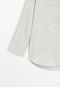 Camisetta a maniche lunghe, grigia chiara a righe, realizzata in tessuto morbido. Presenta un dettaglio con bottone sul petto e un orlo curvo.