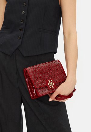 Frau in schwarzem Nadelstreifenanzug, die eine rote, gemusterte Clutch mit goldfarbenem Logo und abnehmbarem Handgelenkriemen hält.