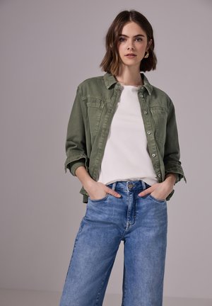 Giacca in denim verde sopra una maglietta bianca, abbinata a jeans a gamba larga azzurri chiari. Presenta chiusura con bottoni e due tasche anteriori.