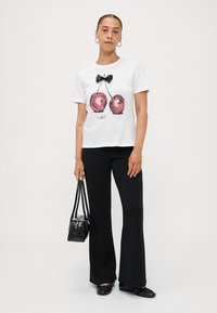 T-shirt en coton blanc avec un graphique de deux cerises rouges avec un nœud noir. Assorti à un pantalon noir évasé et un petit sac à main noir.