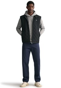 Fekete steppelt mellény, alatta szürke kapucnis pulóverrel, kék nadrággal és krém színű sneakerrel párosítva. A mellény cipzárral záródik és oldalzsebekkel rendelkezik.