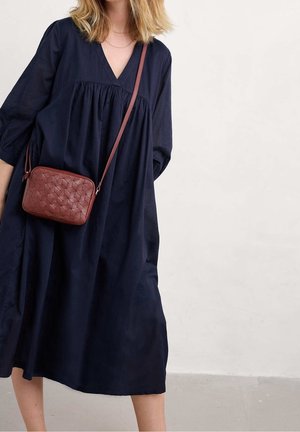 Vrouw draagt een losse marineblauwe jurk en draagt een kleine bruine gestructureerde crossbodytas met een lange band.