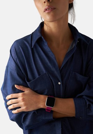 Sort smartwatch med firkantet urskive og lyserød silikonearmbånd, båret på et lyst håndled. Bæreren har en marineblå skjorte med knapper og store lommer.