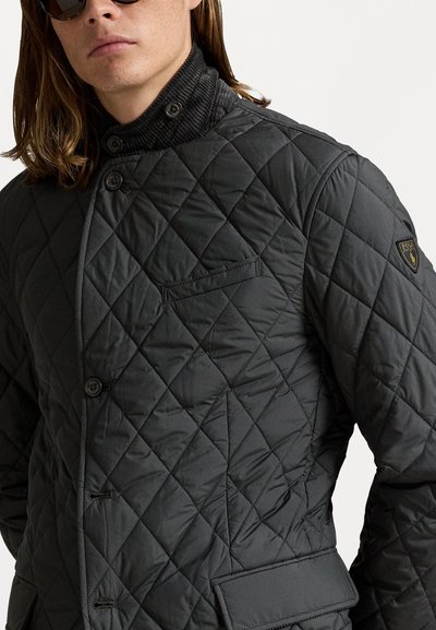 Mann trägt eine schwarze gesteppte Jacke mit Knopfverschluss, Cordkragen und einem Polo Ralph Lauren Logo-Patch am oberen Ärmel.
