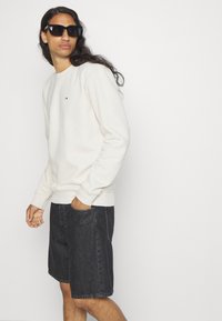 Cremefarbener Sweatshirt mit gerippten Bündchen und kleinem Logodetail. Kombiniert mit dunklen Jeansshorts. Das Model trägt schwarze Sonnenbrille und steht vor einem weißen Hintergrund.