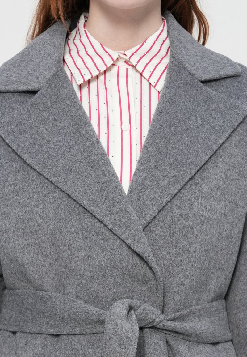 Cappotto di lana grigio con ampi rever legato in vita sopra una camicia bianca con righe verticali rosa e nere e colletto abbottonato.