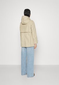 Beige vattentät jacka med huva, två stora sidofickor och dragsko i midjan. Kombinerad med ljusblå vida jeans.