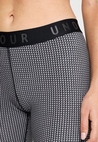 Musta ja harmaa kuvioitu leggingsi, jossa on kiinteä musta elastinen vyötärö, jossa on teksti "UNDER ARMOUR" matta-mustana. Pehmeä, joustava kangas.