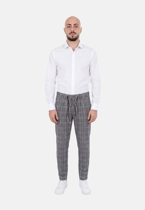 Uomo in piedi di fronte, indossa una camicia bianca con bottoni, pantaloni a quadri grigi con coulisse e scarpe da ginnastica bianche.