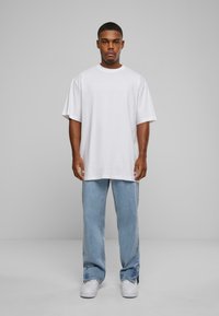Camiseta blanca de gran tamaño con mangas cortas, combinada con jeans de algodón sueltos de color azul claro y zapatillas blancas, de pie contra un fondo gris.
