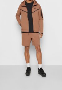 Hnědá mikina na zip s černými detaily, doplněná o hnědé šortky. Černé tričko pod ní. Černé tenisky s bílými ponožkami.