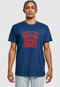 Tamsiai mėlynas medvilninis marškinėliai su ryškiu raudonu „BALLIN“ grafiku ir numeriu „23“ priekyje. Apvalus iškirpimas ir trumpomis rankovėmis.
