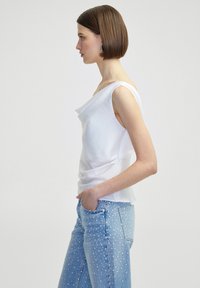 Witte mouwloze top met een gedrapeerd ontwerp, met een gladde textuur. Gecombineerd met lichtblauwe jeans versierd met kleine witte stippen.