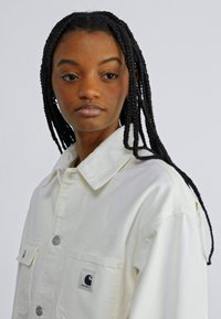 Veste en denim blanc avec un col, poches avant et patch logo circulaire. Présente une coupe décontractée et une texture lisse.