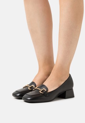 Mocassins en cuir noir avec un bout arrondi, un petit talon bloc et un accent en chaîne dorée sur le dessus du pied. Texture lisse avec un minimum de détails.