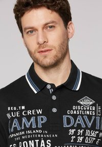 Czarny polo z niebieskimi akcentami, z trzema guzikami i nadrukowanym tekstem szczegółowo opisującym lokalizacje i markę. Kołnierz ma wzór w paski.