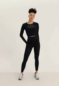 Femme se tenant debout dans un crop top noir à manches longues et des leggings taille haute, portant des chaussures de course argentées, sur un fond clair et uni.
