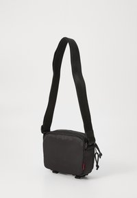 Kompakt sort crossbody-taske med justerbar rem, lynlås hovedrum og tekstureret stofoverflade, stående mod en ensfarvet hvid baggrund.