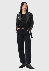 Mujer con chaqueta de cuero negra, camiseta negra con manchas, pantalones vaqueros de talle alto negros con un cinturón ancho y botas negras hasta el tobillo, de pie con las manos en los bolsillos.
