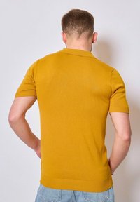 Polo à manches courtes en jaune moutarde, fabriqué dans un tissu doux et extensible. Doté d'un col plat et de poignets côtelés. Design minimaliste sans motifs visibles.