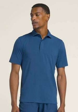 Uomo che indossa una polo blu a maniche corte e pantaloni blu abbinati, che guarda leggermente verso sinistra su uno sfondo neutro.