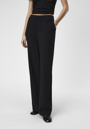 Zwarte wijde broek met een gladde textuur, hoge taille, en zijzakken, gecombineerd met glanzende zwarte schoenen.