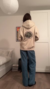 Béžová mikina s kapucí a kruhovým motivem na zádech, doplněná světle modrými džínami s širokými nohavicemi a černou obuví, stojící uvnitř.