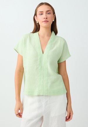 Femme aux longs cheveux bruns portant des boucles d’oreilles crème, un chemisier vert clair à manches courtes et col en V, et un pantalon blanc, debout devant un fond uni.