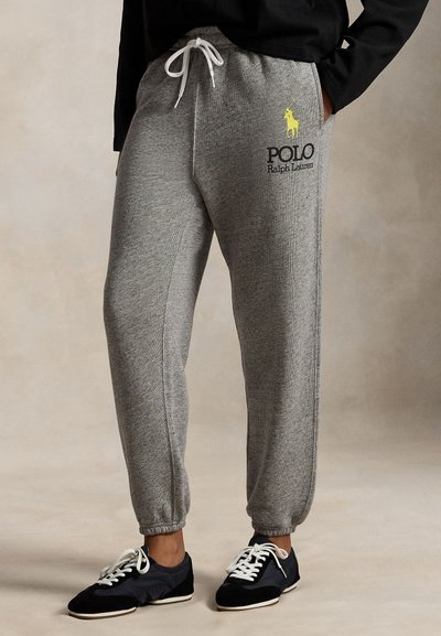 Grå sweatpants med elastiske manchetter og snøre i taljen. Har gul "Polo Ralph Lauren"-logo på venstre ben. Et par sorte sko er synlige.