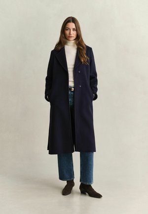 Femme portant un long manteau foncé, un col roulé blanc, un jean bleu et des bottines foncées, debout devant un fond clair uni.