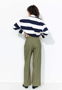 Chemise rayée oversize bleu marine et blanche à épaules tombantes, associée à un pantalon taille haute vert olive. Chevilles noires cloutées visibles.