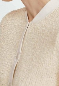 Beżowa kurtka o fakturze z okrągłym dekoltem, z zapięciem na zamek z przodu oraz gładkimi, jasnymi guzikami wzdłuż zapięcia.