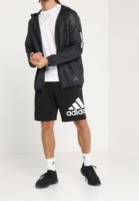 Czarna lekkie kurtka z kapturem, białe paski po bokach, białe logo Adidasa. Biała koszulka, czarne szorty z dużym białym logo Adidasa, czarne sneakersy.