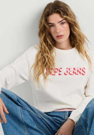 Noor naine pikkade laineliste juustega, kellel on seljas valge dressipluus punase "PEPE JEANS" kirjaga, istub vabalt siniste teksadega ühevärvilise taustaga.