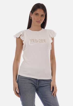 Donna che indossa una maglietta bianca con maniche arricciate e la scritta "YES ZEE", abbinata a jeans blu, in piedi davanti a uno sfondo neutro.