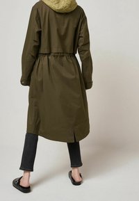 Cappotto lungo verde oliva con cappuccio imbottito, vita stretta e fessure laterali. Indossato sopra jeans scuri e zoccoli neri. Materiale liscio e resistente all'acqua.