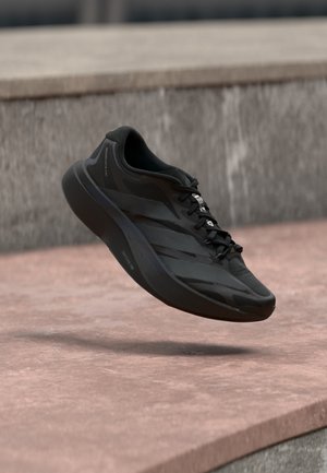 Chaussure de course moderne noire en suspension au-dessus de marches en béton rose avec un fond en pierre grise, mettant en valeur sa semelle et les détails de son design.
