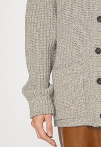 Ljusgrå stickad cardigan med en strukturerad mönster, som har stora knappar, ribbade ärmslut och en framtill ficka.