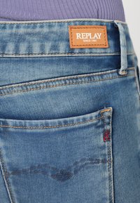 Ανοιχτό μπλε τζιν παντελόνι με υφή, διαθέτοντας μία καφέ δερμάτινη ετικέτα με την ένδειξη "REPLAY" και διακοσμητική ραφή στις πίσω τσέπες.