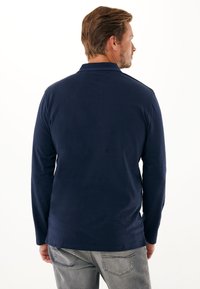 Polo a maniche lunghe navy in tessuto morbido; presenta un colletto classico e un orlo dritto, indossato con jeans grigi chiari.