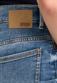 Blauwe denim jeans met gele stiksels, voorzien van een bruine lederen patch met het label "DRYKORN" op de achterkant van de taille.