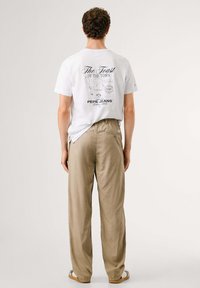 Weißes T-Shirt mit einem grafischen Druck auf dem Rücken und beigen, locker sitzenden Hosen. Das T-Shirt zeigt Text und Cartoon-Designs.