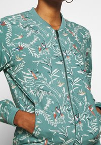 Veste bomber bleu sarcelle avec un motif floral et des oiseaux, fermeture éclair, poignets côtelés, coupe décontractée, confectionnée dans un tissu doux et texturé.