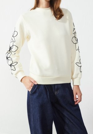 Kvinde iført en cremefarvet sweatshirt med sorte blomsterlinjedesigns på begge ærmer og mørkeblå vidde bukser.