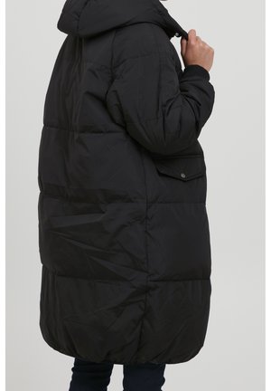 Person, der einen langen schwarzen gesteppter Mantel mit Kapuze, Seitentaschen mit Klappen und gerippten Bündchen trägt und vor einem schlichten, hellen Hintergrund steht.