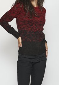 Gestrickter Pullover mit einem Farbverlauf von Rot zu Schwarz, mit einem strukturierten Muster sowie gerippten Bündchen und Saum. Kombiniert mit schwarzen Jeans, die mit Akzenten verziert sind.