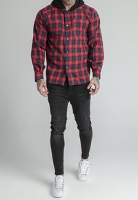 Camicia flanella a quadri rossi e blu con bottoni, una tasca e cappuccio nero, abbinata a jeans skinny neri e sneakers bianche.