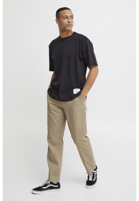Schwarzes Kurzarm-T-Shirt mit lockerer Passform, kombiniert mit beigen Hosen und schwarzen Sneakers, mit einem sichtbaren Markenschild an der Seite des Shirts.