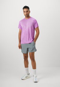 Lila Sport-T-Shirt mit gestrickter Textur, kombiniert mit grauen Shorts. Die Schuhe sind weiß mit schwarzen Akzenten. Neutraler Hintergrund.